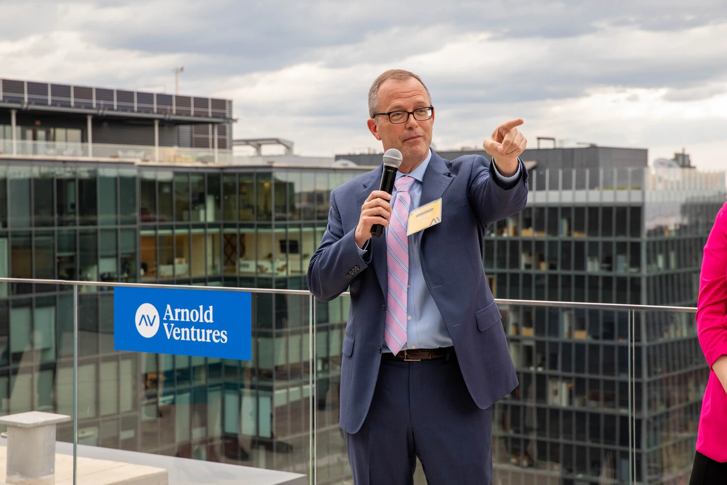 A man in a suit holds a microphone and gestures while speaking on a rooftop with city buildings in the background. A sign reads Arnold Ventures.