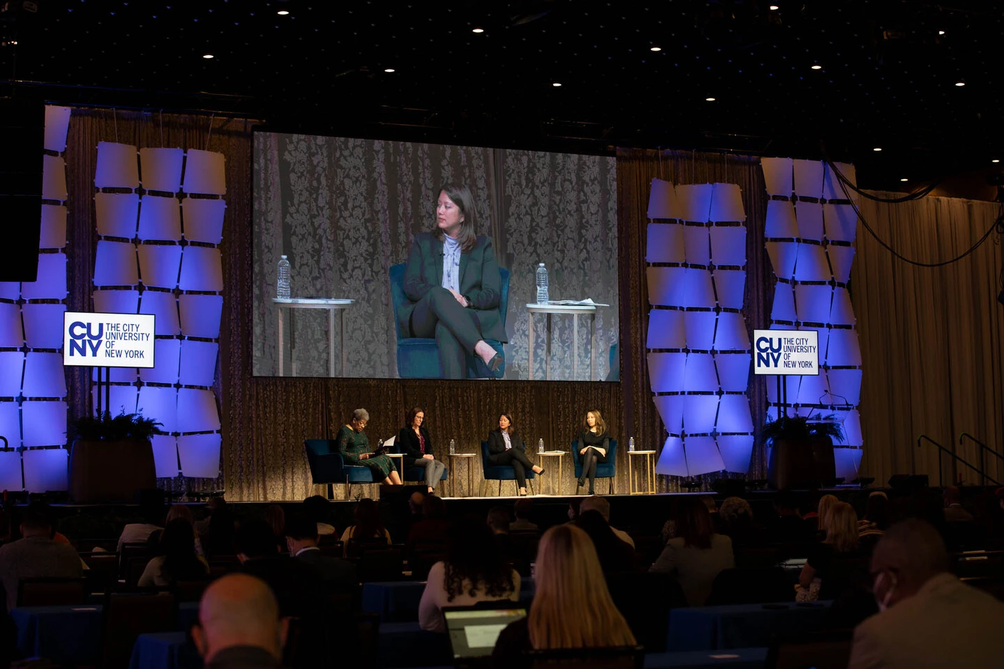 Panel discussion at CUNY with four women seated on stage, a large screen displaying one speaker. Audience watches attentively in a dimly lit room