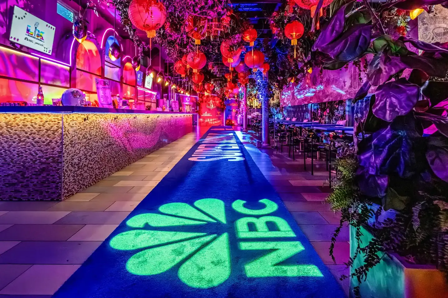 Vibrant bar with pink, purple, and blue lighting, red lanterns, and lush greenery. NBC logo projected on a blue carpet, creating a lively ambiance